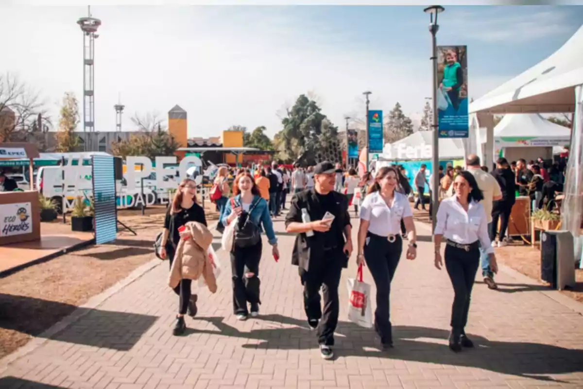 Personas caminando por un paseo al aire libre durante un evento o feria con puestos y carpas a los costados en un día soleado