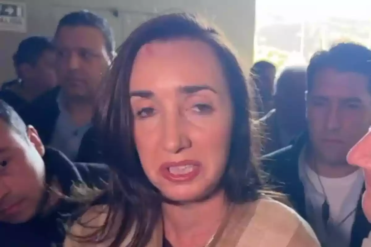 Mujer con expresión de dolor y una herida en la frente rodeada de varias personas en un ambiente interior