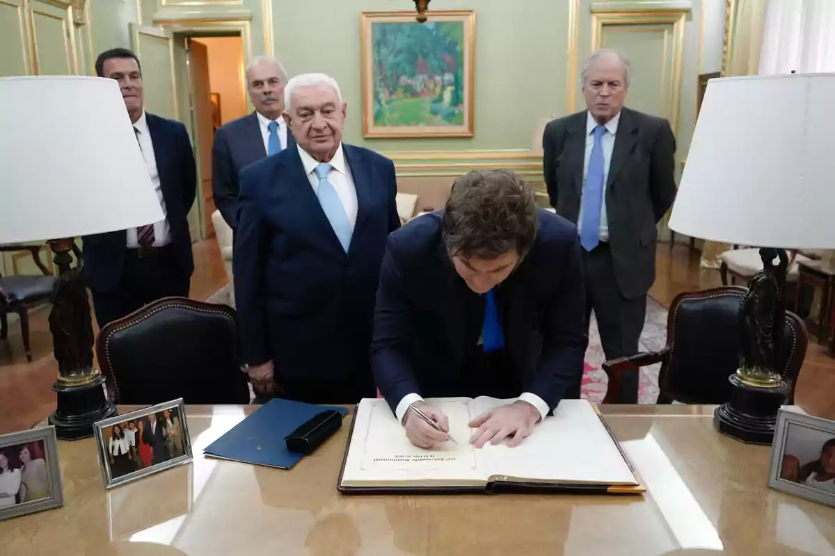 Un hombre firma un libro grande sobre un escritorio mientras cuatro hombres de traje lo observan en una oficina elegante decorada con cuadros, lámparas y fotos familiares.