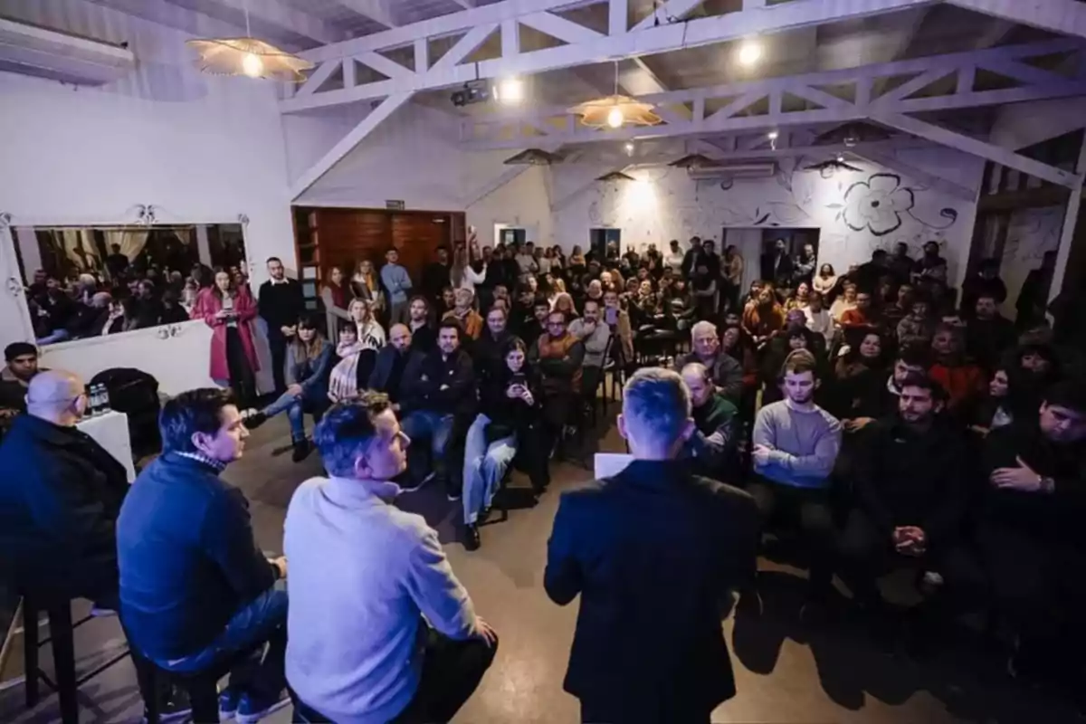 Un grupo de personas asiste a una charla o conferencia en un salón iluminado con techos altos y decoración artística en las paredes, mientras varios oradores están sentados al frente dialogando con el público. Un grupo de personas asiste a una charla o conferencia en un salón iluminado con techos altos y decoración artística en las paredes, mientras varios oradores están sentados al frente dialogando con el público.
