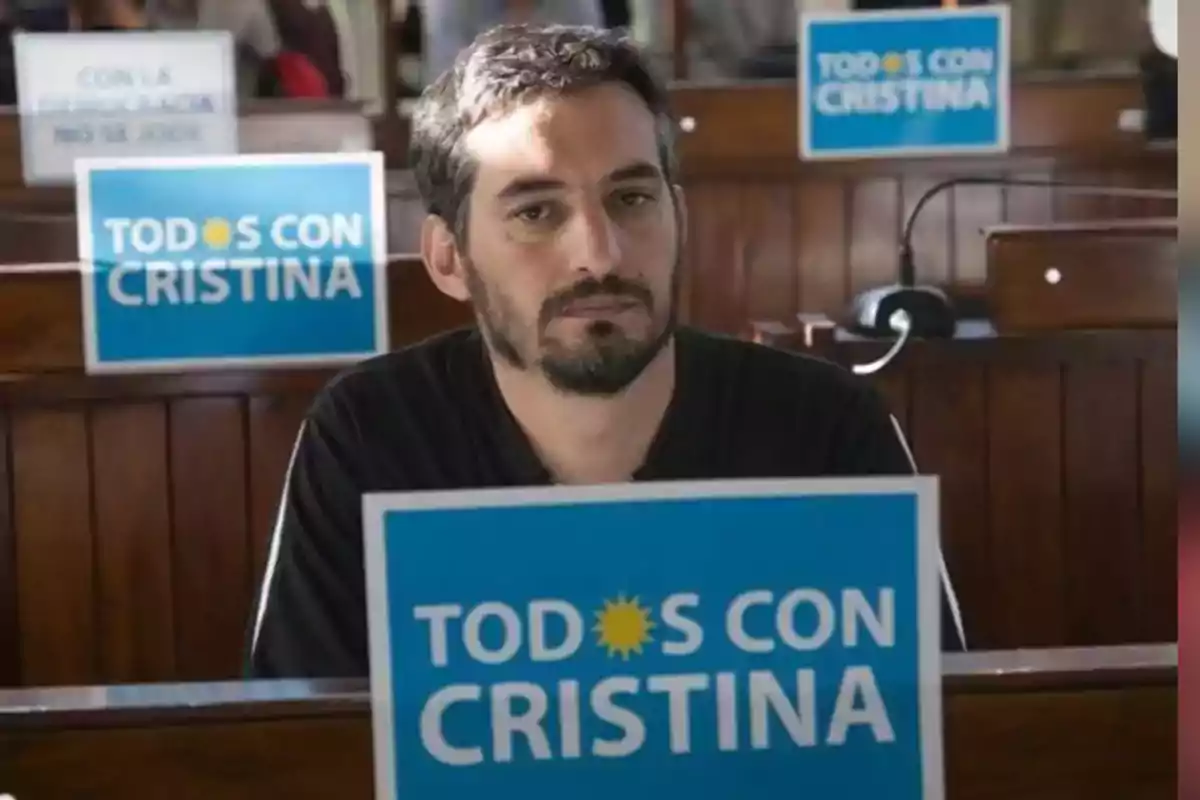 Hombre sentado en un recinto con carteles azules que dicen TODOS CON CRISTINA Hombre sentado en un recinto con carteles azules que dicen TODOS CON CRISTINA