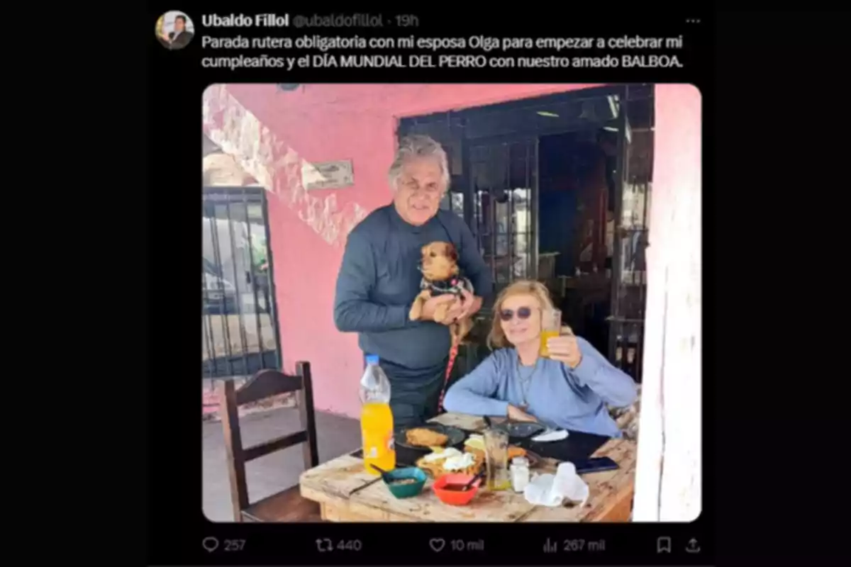 Una pareja mayor disfruta de una comida al aire libre en una mesa de madera, la mujer sostiene un vaso con bebida y el hombre está de pie sosteniendo un perro pequeño, ambos sonríen y hay platos y una botella de gaseosa sobre la mesa Una pareja mayor disfruta de una comida al aire libre en una mesa de madera, la mujer sostiene un vaso con bebida y el hombre está de pie sosteniendo un perro pequeño, ambos sonríen y hay platos y una botella de gaseosa sobre la mesa