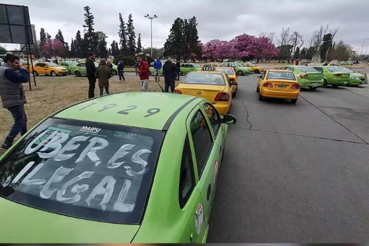 Varios taxis verdes y amarillos están estacionados en una calle mientras un grupo de personas conversa cerca, en el parabrisas trasero de uno de los autos se lee un mensaje que dice Uber es ilegal. Varios taxis verdes y amarillos están estacionados en una calle mientras un grupo de personas conversa cerca, en el parabrisas trasero de uno de los autos se lee un mensaje que dice Uber es ilegal.
