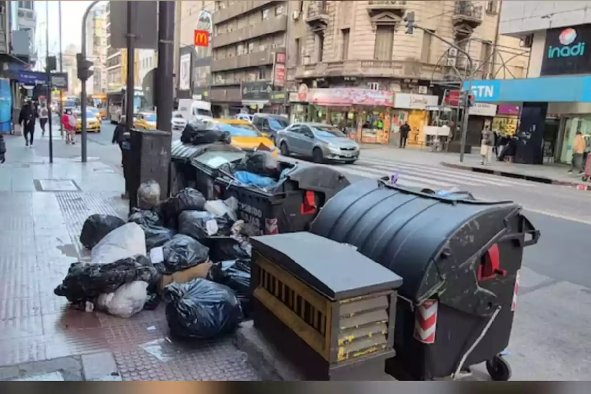 Bolsas de basura acumuladas en la acera junto a contenedores en una calle urbana con tráfico y comercios al fondo Bolsas de basura acumuladas en la acera junto a contenedores en una calle urbana con tráfico y comercios al fondo