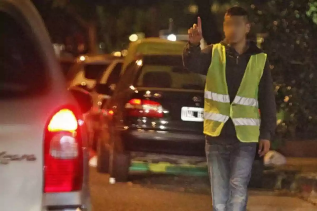 Un hombre con chaleco reflectante está de pie en la calle de noche levantando un dedo mientras varios autos están estacionados detrás de él. Un hombre con chaleco reflectante está de pie en la calle de noche levantando un dedo mientras varios autos están estacionados detrás de él.