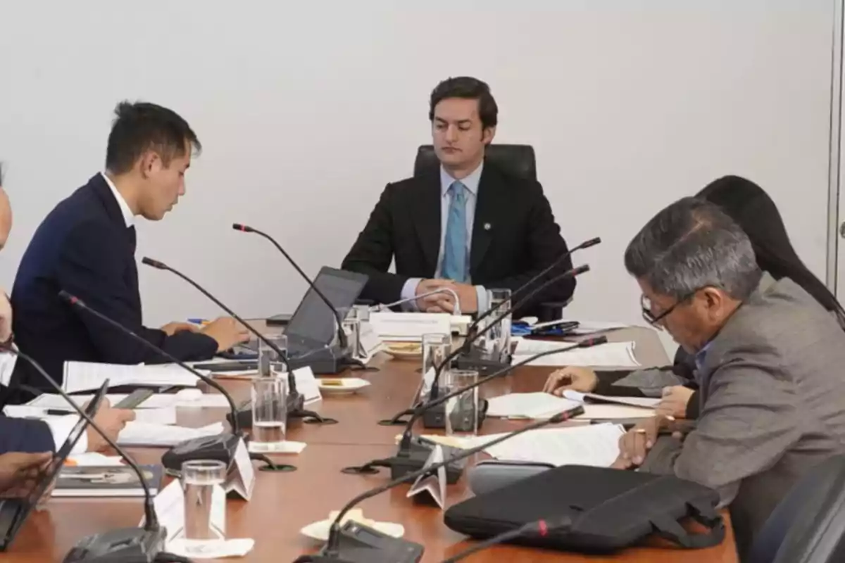 Varias personas con traje están sentadas alrededor de una mesa de reuniones con micrófonos, laptops y documentos, participando en una junta de trabajo en una oficina.