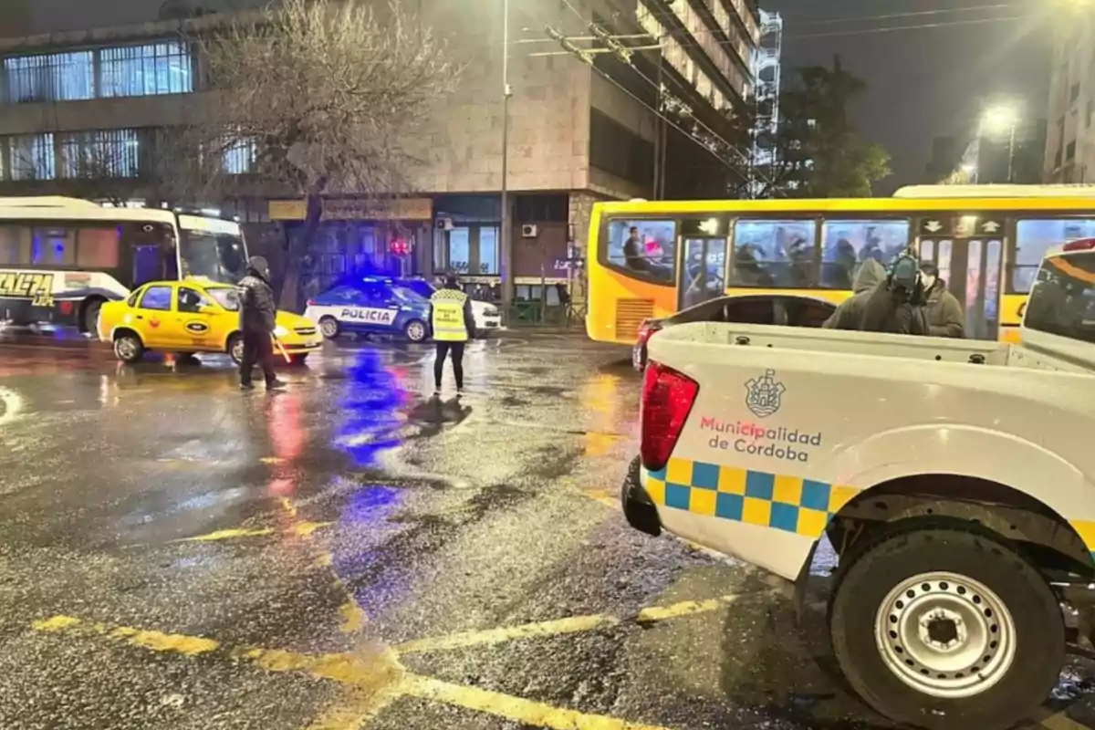 Traffic officers and police coordinate traffic on a rain-soaked street, with official vehicles, a taxi, and a bus present at an urban intersection in Córdoba.