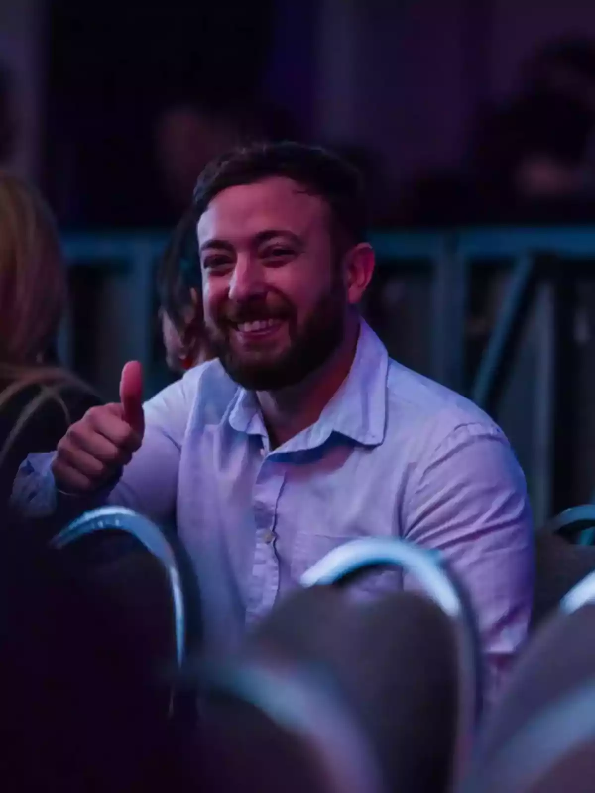 Hombre con barba y camisa clara sentado en una silla mostrando una señal de pulgar arriba y sonriendo en un ambiente interior iluminado con luces tenues. Hombre con barba y camisa clara sentado en una silla mostrando una señal de pulgar arriba y sonriendo en un ambiente interior iluminado con luces tenues.