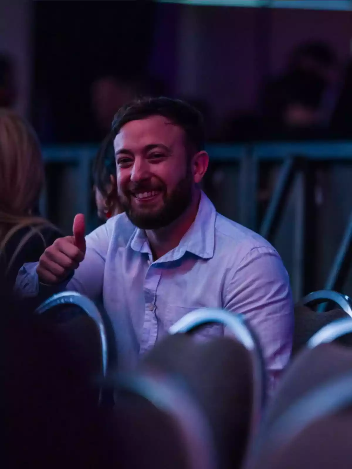 Hombre joven con barba y camisa clara sentado entre sillas en un evento sonríe y hace un gesto de pulgar arriba Hombre joven con barba y camisa clara sentado entre sillas en un evento sonríe y hace un gesto de pulgar arriba