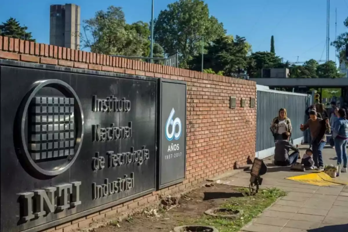 Main entrance of the National Institute of Industrial Technology with a 60th anniversary commemorative sign and several people on the sidewalk next to a brick wall. Main entrance of the National Institute of Industrial Technology with a 60th anniversary commemorative sign and several people on the sidewalk next to a brick wall.