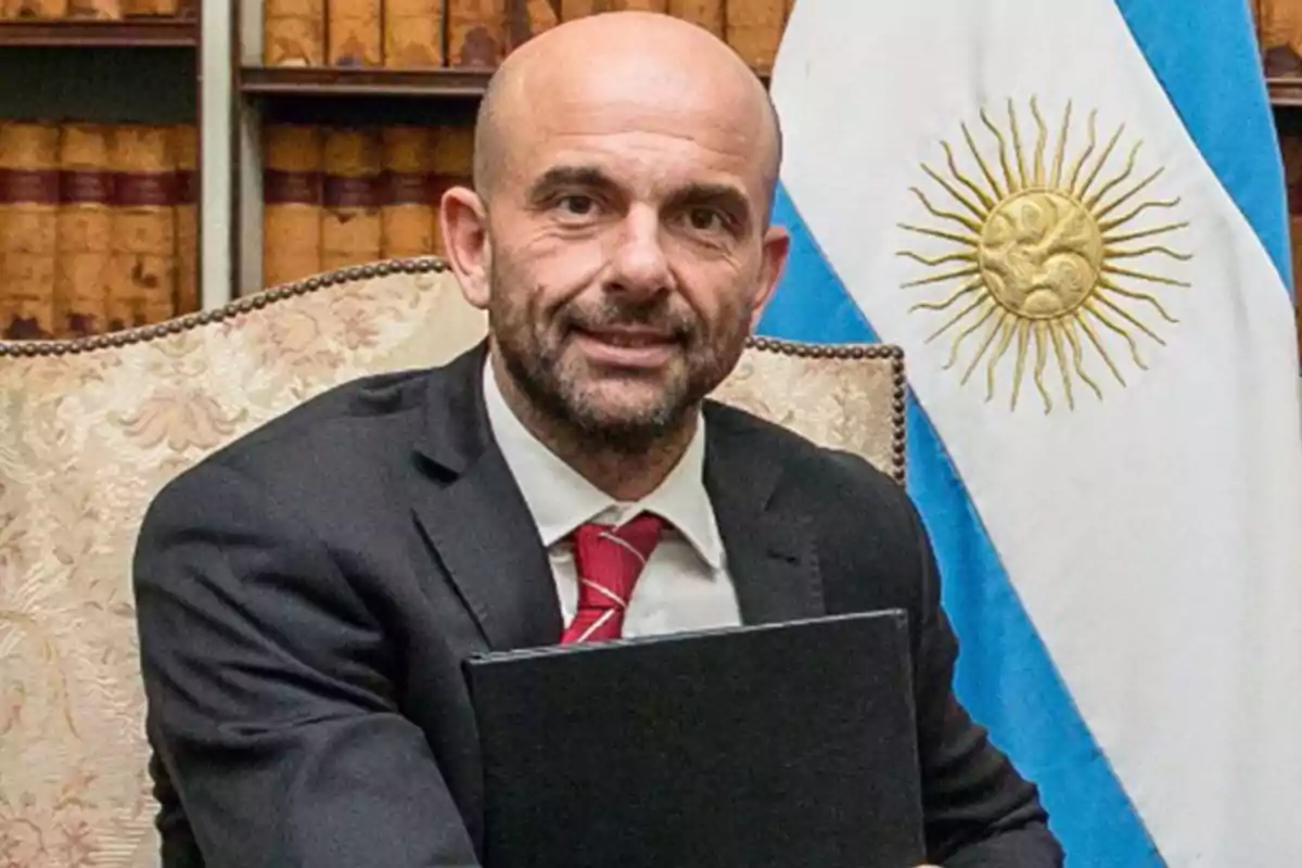 Hombre calvo con barba y traje oscuro sosteniendo una carpeta negra, sentado frente a una bandera de Argentina y una estantería con libros antiguos. Hombre calvo con barba y traje oscuro sosteniendo una carpeta negra, sentado frente a una bandera de Argentina y una estantería con libros antiguos.