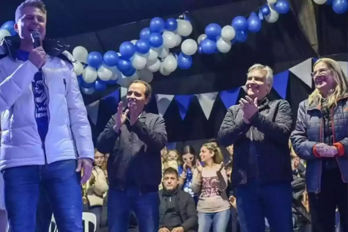 Un hombre con micrófono habla en un escenario decorado con globos y banderines azules y blancos mientras otras personas aplauden y sonríen a su lado Un hombre con micrófono habla en un escenario decorado con globos y banderines azules y blancos mientras otras personas aplauden y sonríen a su lado