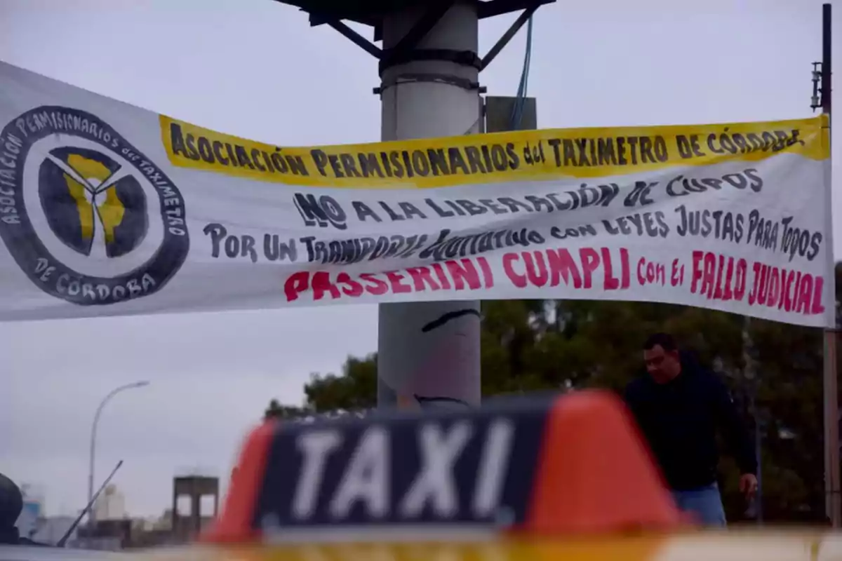 Cartel de la Asociación Permisionarios del Taxímetro de Córdoba colgado en la vía pública con mensajes de protesta y un taxi en primer plano desenfocado Cartel de la Asociación Permisionarios del Taxímetro de Córdoba colgado en la vía pública con mensajes de protesta y un taxi en primer plano desenfocado