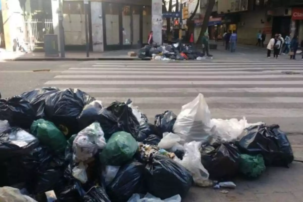Bolsas de basura acumuladas en una esquina de una calle urbana con personas caminando al fondo Bolsas de basura acumuladas en una esquina de una calle urbana con personas caminando al fondo
