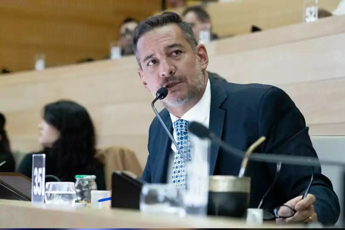Hombre de traje hablando en un micrófono durante una sesión en un auditorio con otras personas al fondo y objetos sobre la mesa Hombre de traje hablando en un micrófono durante una sesión en un auditorio con otras personas al fondo y objetos sobre la mesa