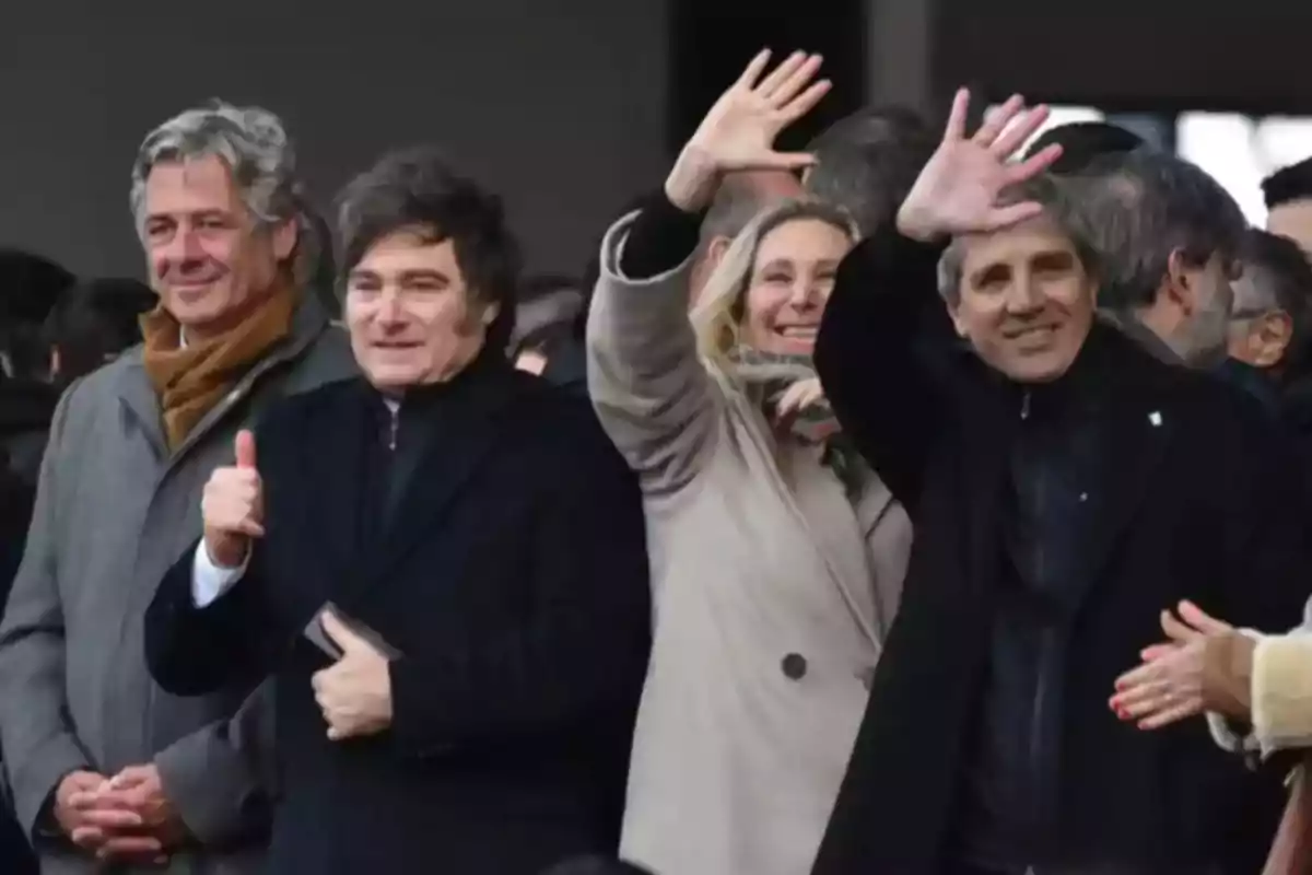 Cuatro personas de pie en un evento al aire libre, dos de ellas saludan con la mano y una muestra el pulgar hacia arriba, todos visten ropa de abrigo y parecen sonrientes. Cuatro personas de pie en un evento al aire libre, dos de ellas saludan con la mano y una muestra el pulgar hacia arriba, todos visten ropa de abrigo y parecen sonrientes.