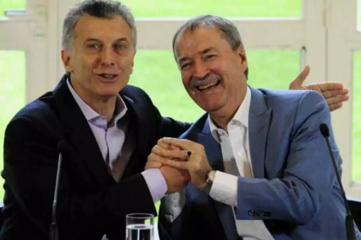 Two men in suits smile and greet each other enthusiastically while sitting in front of microphones. Two men in suits smile and greet each other enthusiastically while sitting in front of microphones.