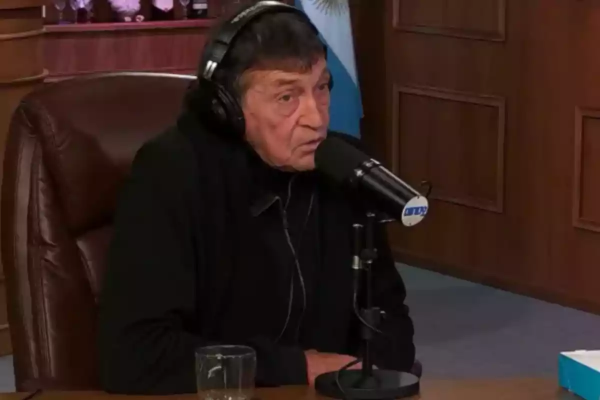 Hombre mayor con auriculares hablando frente a un micrófono en un estudio de radio con una bandera de Argentina de fondo