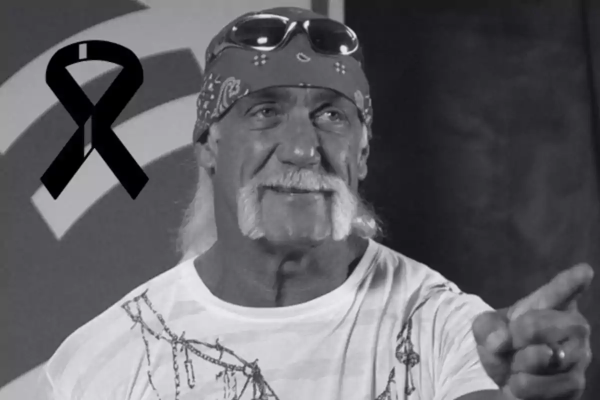 Long-haired man with a mustache, wearing a bandana and glasses on his head, pointing forward, black and white image with a black mourning bow in the corner