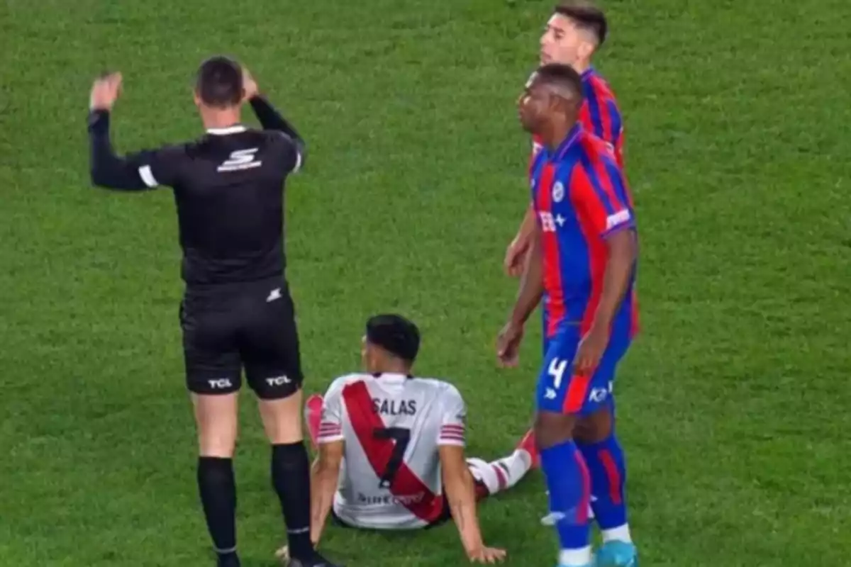 Un árbitro de fútbol señala una jugada mientras un jugador con uniforme blanco y rojo está sentado en el césped y dos jugadores con uniforme azul y rojo observan la situación.