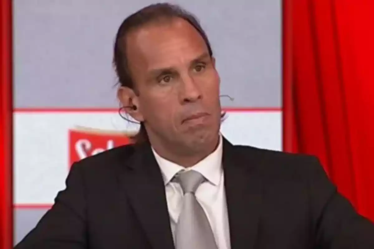 Hombre de cabello largo vestido con traje oscuro y corbata gris sentado en un estudio de televisión con fondo rojo y gris Hombre de cabello largo vestido con traje oscuro y corbata gris sentado en un estudio de televisión con fondo rojo y gris
