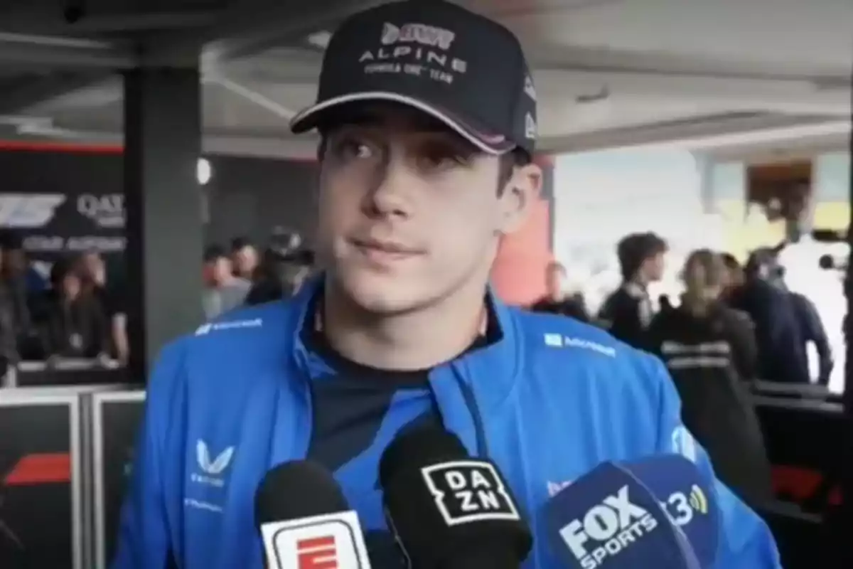 Motorsport driver wearing a cap and blue jacket giving an interview in front of several microphones from different media outlets in a race paddock.