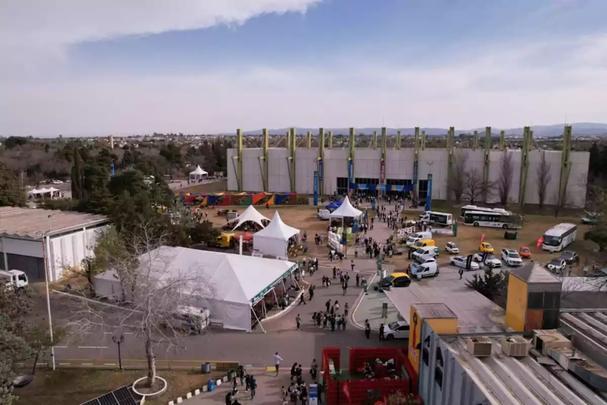 Vista aérea de un recinto ferial con varias carpas blancas, personas caminando, autos y autobuses estacionados, y un edificio grande al fondo rodeado de árboles