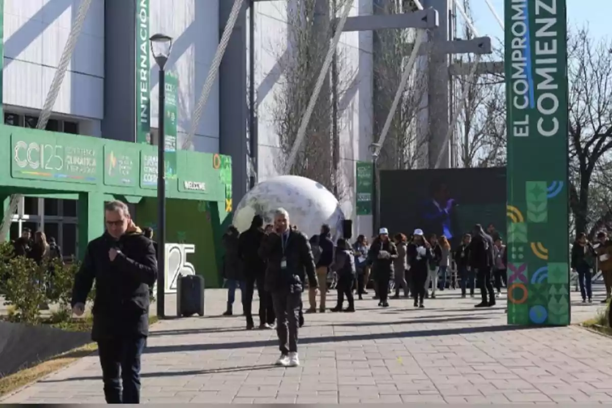 Personas caminando y conversando en la entrada de un evento sobre cambio climático con carteles verdes y una esfera decorativa