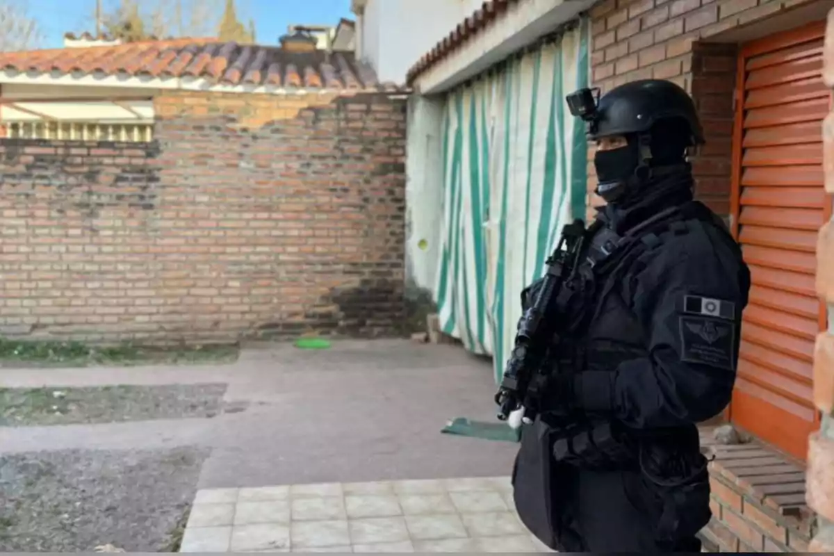 Un agente de policía fuertemente armado y vestido con uniforme táctico negro se encuentra de pie en el patio de una casa junto a una pared de ladrillos y una puerta enrollable naranja