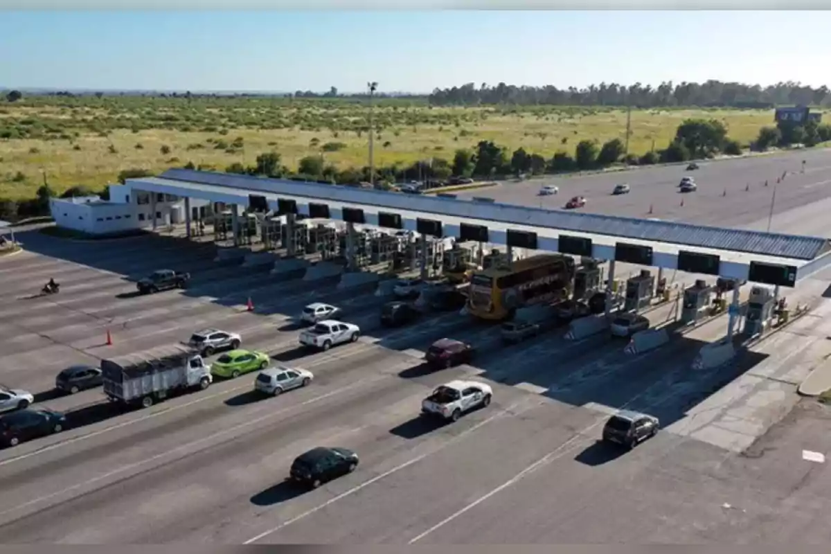 Caseta de peaje en una autopista con varios vehículos haciendo fila para pagar, rodeada de áreas verdes y cielo despejado