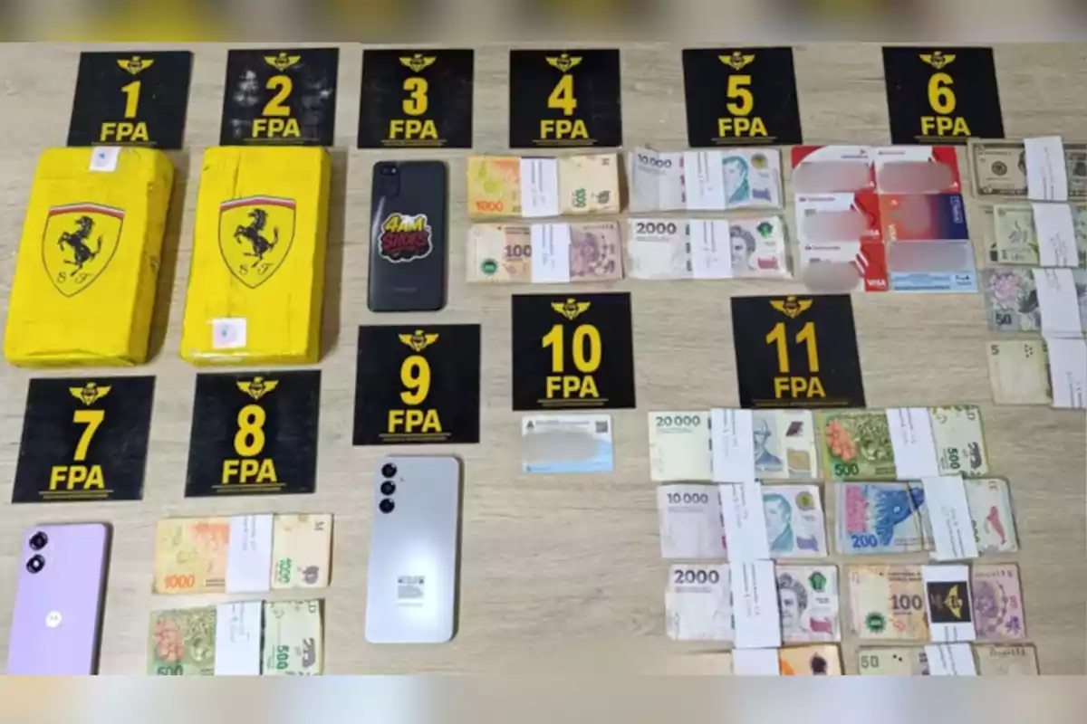 Evidencia incautada organizada sobre una mesa que incluye dos paquetes amarillos con logotipo de Ferrari, varios fajos de billetes de diferentes denominaciones y monedas, tres teléfonos móviles, varias tarjetas bancarias y documentos, todos identificados con números de la FPA