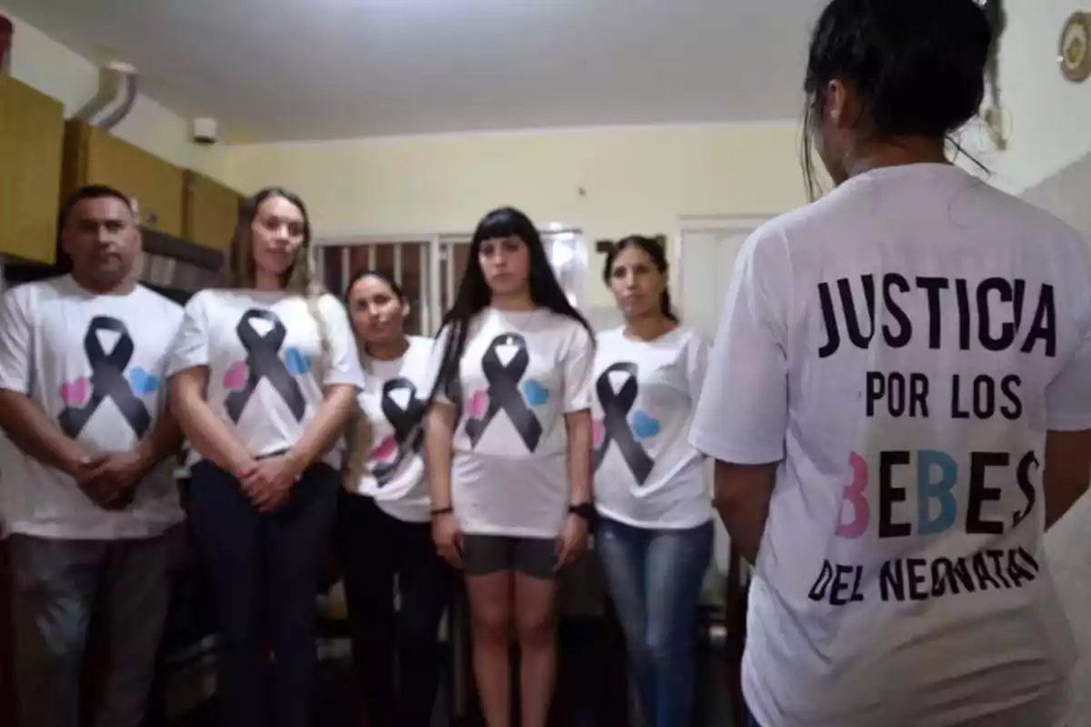 Grupo de personas con camisetas blancas que tienen un lazo negro y una mujer de espaldas con una camiseta que dice justicia por los bebes del neonatológico