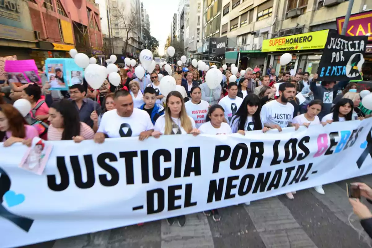 Manifestación en la calle con personas sosteniendo un cartel que pide justicia por los bebés del área neonatal, acompañados de globos blancos y pancartas.