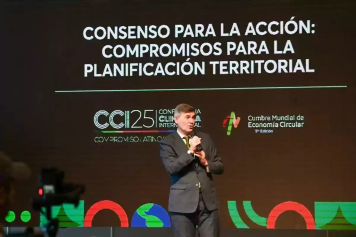 Hombre de traje hablando en un escenario durante un evento sobre planificación territorial y economía circular con un fondo que muestra el título del evento y logotipos relacionados