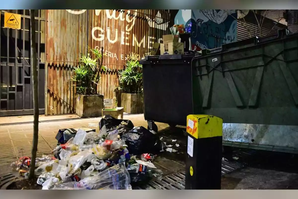 Basura acumulada en la calle junto a un contenedor desbordado frente a una galería comercial por la noche