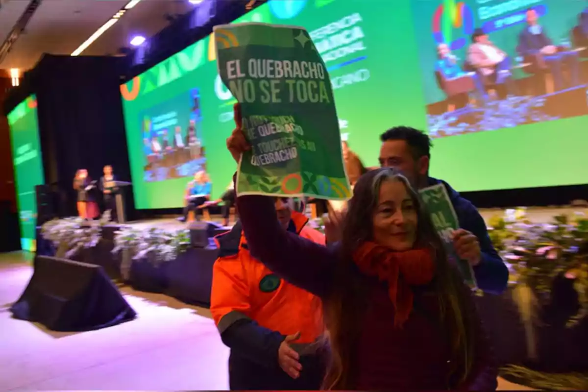 Mujer con bufanda roja sostiene un cartel verde que dice El quebracho no se toca en un evento con escenario y pantallas al fondo