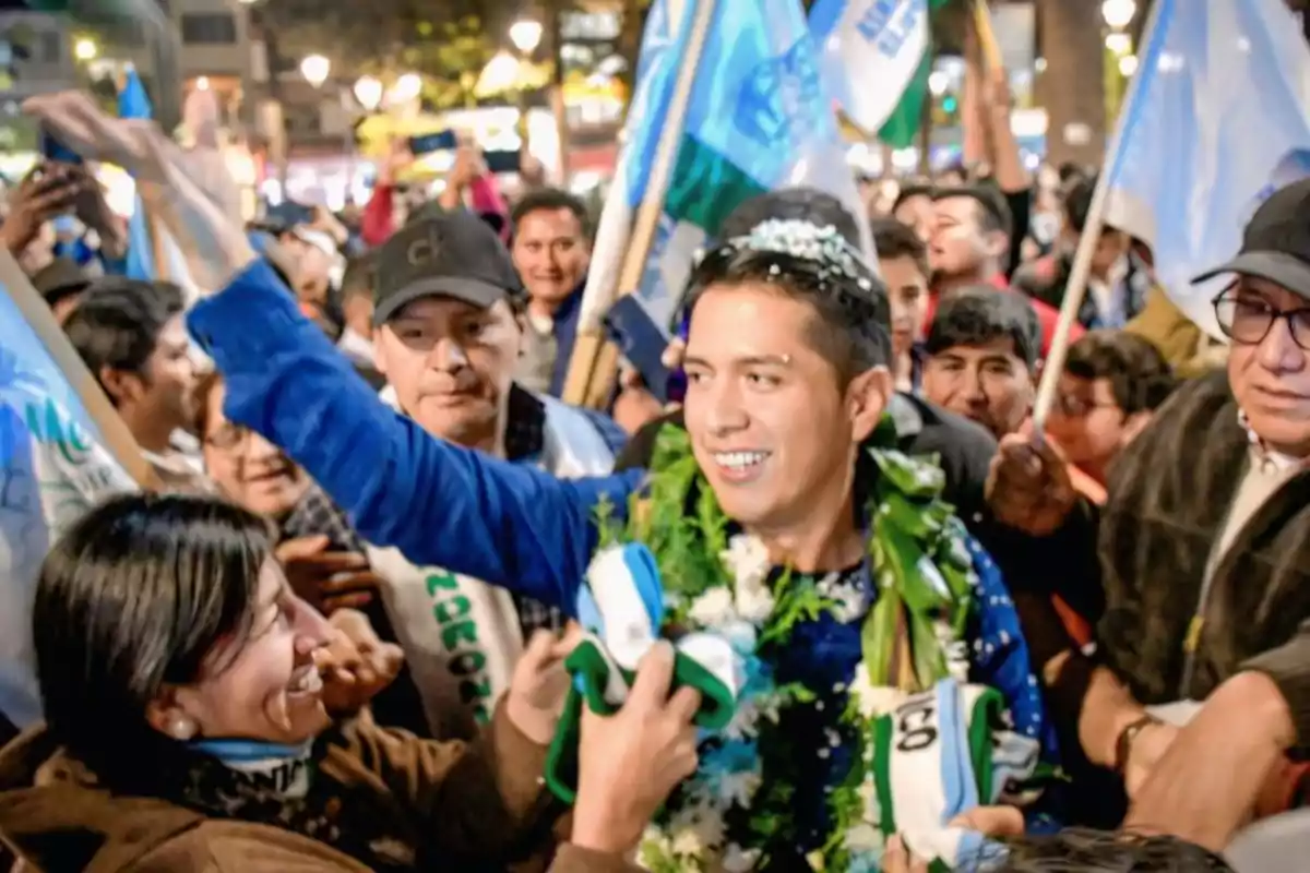 Un hombre sonriente con guirnaldas de flores es rodeado por una multitud de personas que celebran y ondean banderas en un evento al aire libre por la noche Un hombre sonriente con guirnaldas de flores es rodeado por una multitud de personas que celebran y ondean banderas en un evento al aire libre por la noche