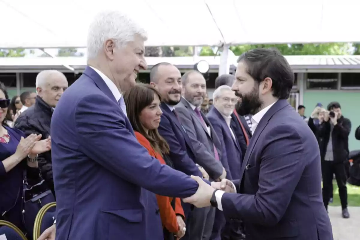 Two men in suits shake hands at an outdoor formal event while several people watch and applaud in the background.