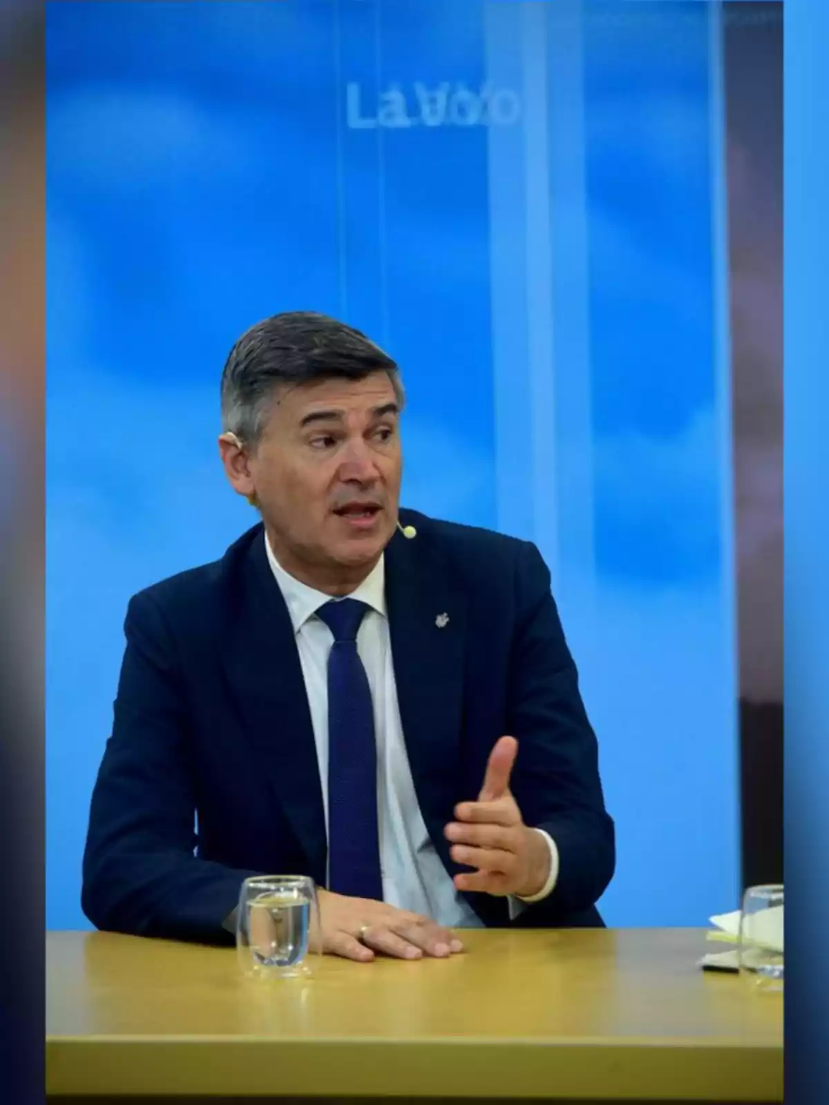 Hombre de cabello corto y canoso con traje azul marino y corbata azul hablando mientras gesticula con la mano sentado en una mesa con vasos de agua y fondo azul Hombre de cabello corto y canoso con traje azul marino y corbata azul hablando mientras gesticula con la mano sentado en una mesa con vasos de agua y fondo azul