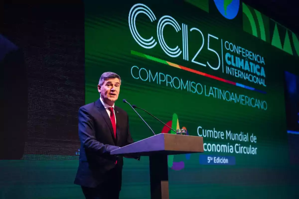 Hombre de traje y corbata roja hablando en un podio durante una conferencia climática internacional con un fondo verde que muestra el texto Compromiso Latinoamericano y Cumbre Mundial de Economía Circular Hombre de traje y corbata roja hablando en un podio durante una conferencia climática internacional con un fondo verde que muestra el texto Compromiso Latinoamericano y Cumbre Mundial de Economía Circular