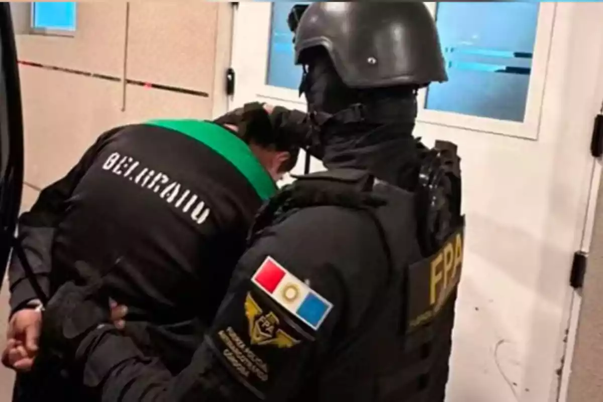 Un agente de la fuerza policial arresta a una persona con un uniforme que tiene la palabra Belgrano en la espalda dentro de un edificio. Un agente de la fuerza policial arresta a una persona con un uniforme que tiene la palabra Belgrano en la espalda dentro de un edificio.