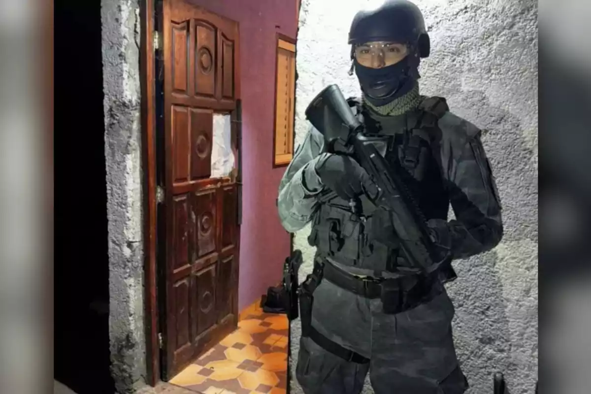 Policía fuertemente armado con uniforme táctico y casco de protección parado junto a una puerta de madera abierta en una casa de paredes moradas y piso de mosaico Policía fuertemente armado con uniforme táctico y casco de protección parado junto a una puerta de madera abierta en una casa de paredes moradas y piso de mosaico