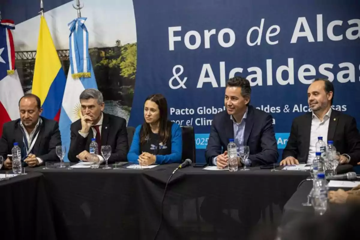 Five people sitting at a table during a mayors’ forum, with flags from several countries in the background and water bottles on the table. Five people sitting at a table during a mayors’ forum, with flags from several countries in the background and water bottles on the table.
