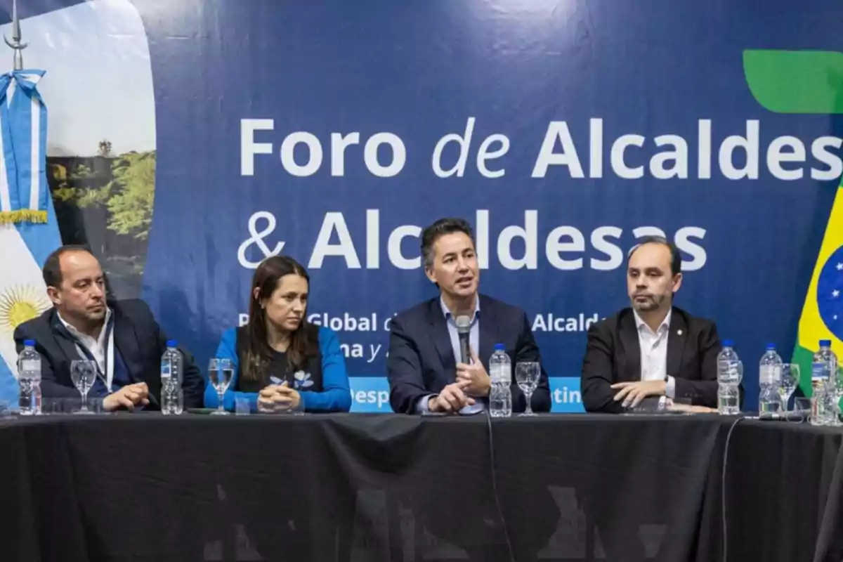 Four people seated at a table during a mayors’ forum with Argentine and Brazilian flags in the background Four people seated at a table during a mayors’ forum with Argentine and Brazilian flags in the background