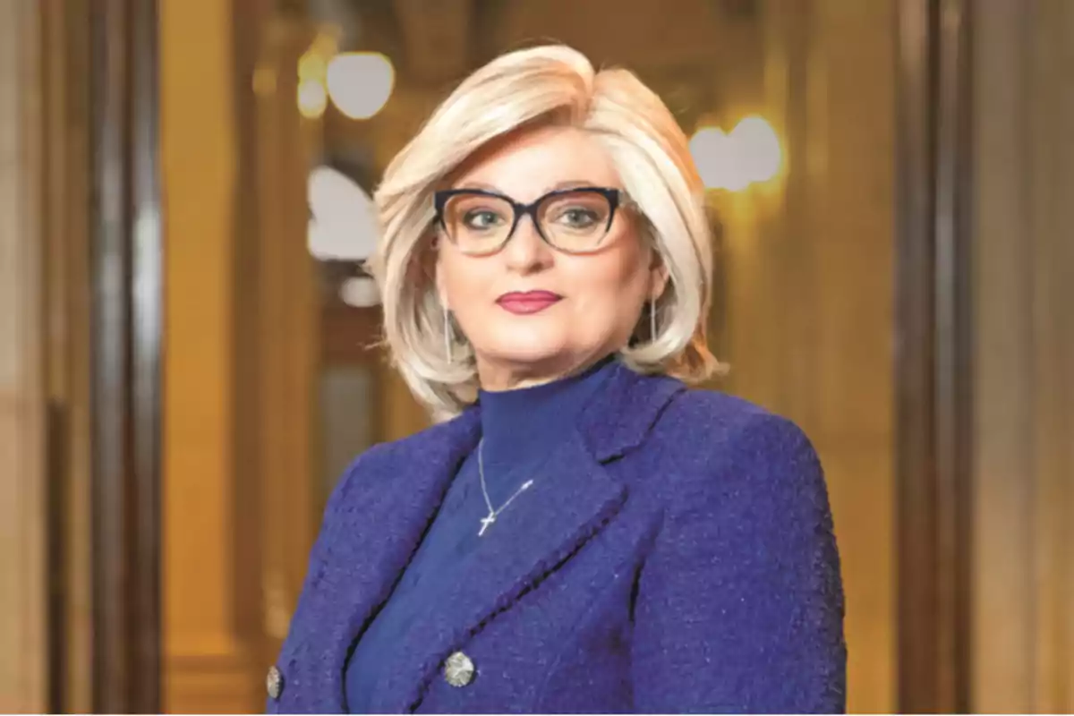 Short-haired blonde woman with glasses and a blue blazer posing in an elegant, well-lit setting Short-haired blonde woman with glasses and a blue blazer posing in an elegant, well-lit setting