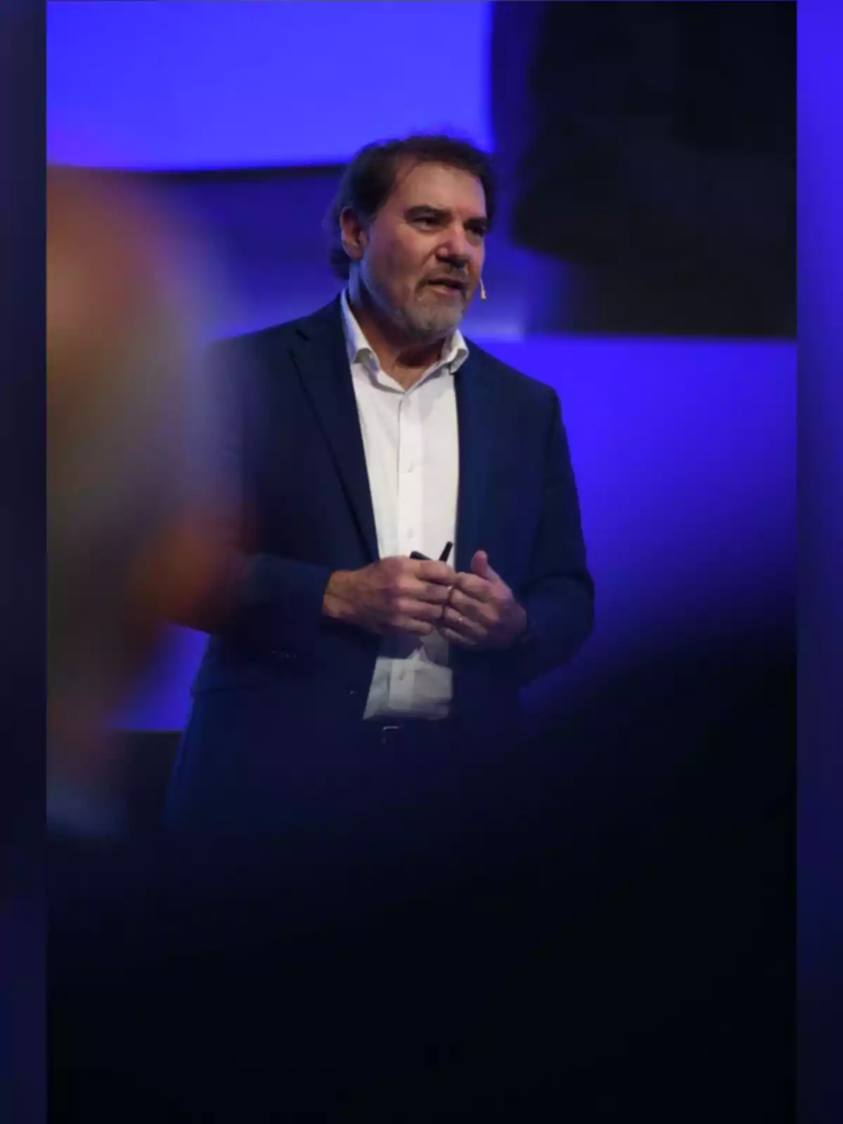 Hombre de mediana edad con barba y cabello castaño usando un micrófono de diadema y traje azul oscuro mientras habla en un escenario iluminado con luces azules Hombre de mediana edad con barba y cabello castaño usando un micrófono de diadema y traje azul oscuro mientras habla en un escenario iluminado con luces azules