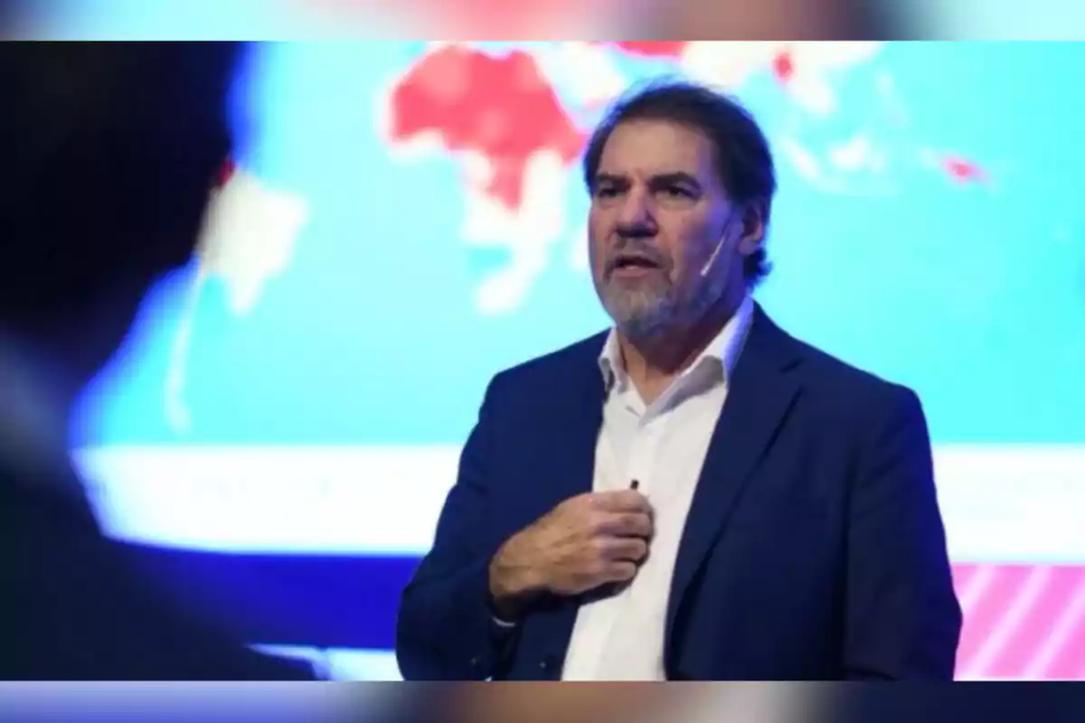 Hombre de mediana edad con barba y cabello oscuro usando un micrófono de diadema y traje azul mientras habla frente a una pantalla con un mapa de fondo borroso Hombre de mediana edad con barba y cabello oscuro usando un micrófono de diadema y traje azul mientras habla frente a una pantalla con un mapa de fondo borroso