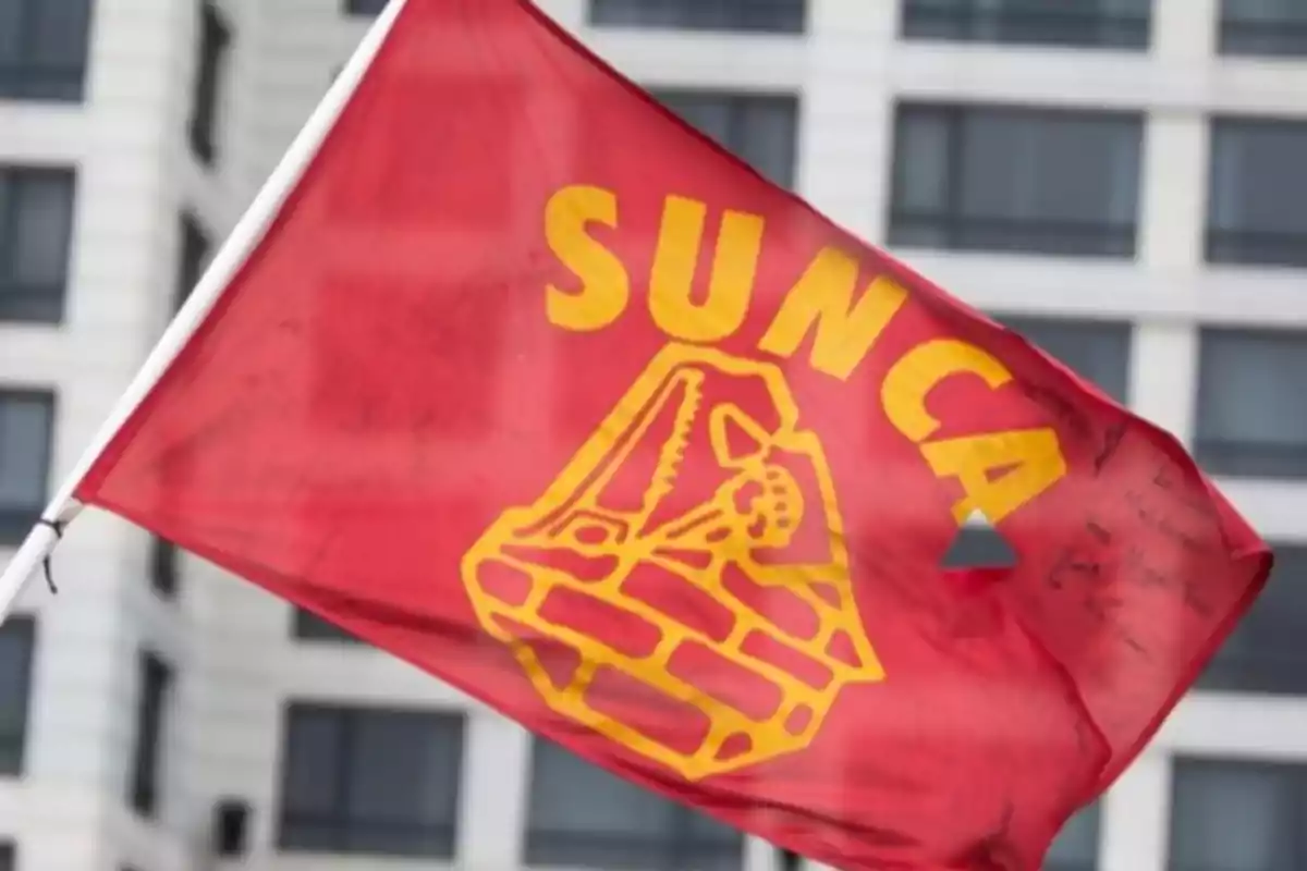 Bandera roja con el logo y las letras amarillas de SUNCA ondeando frente a un edificio Bandera roja con el logo y las letras amarillas de SUNCA ondeando frente a un edificio