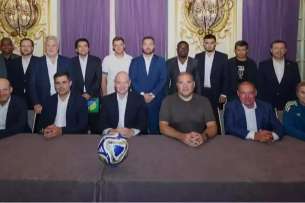 Un grupo de personas vestidas de manera formal posan detrás de una mesa con un balón de fútbol en el centro, en una sala decorada con cortinas moradas y detalles dorados en la pared. Un grupo de personas vestidas de manera formal posan detrás de una mesa con un balón de fútbol en el centro, en una sala decorada con cortinas moradas y detalles dorados en la pared.