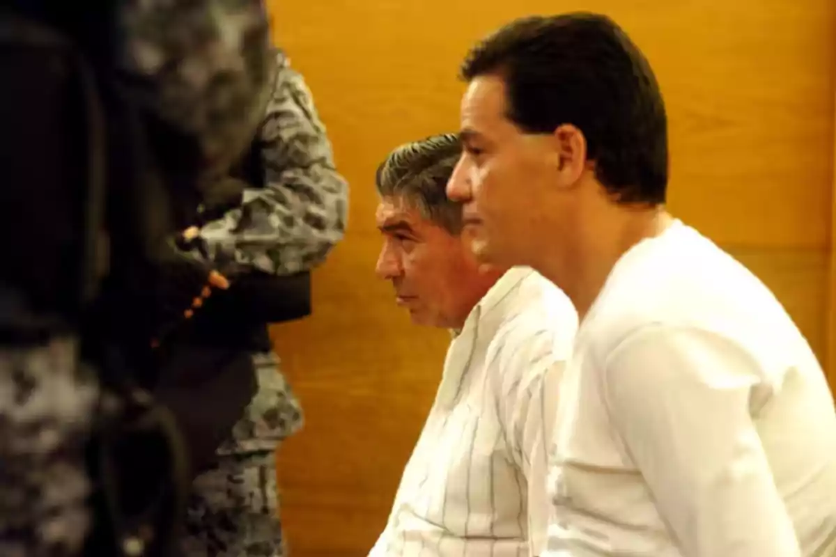 Two men sitting with serious expressions next to a guard dressed in tactical uniform in an indoor setting with wood-colored walls Two men sitting with serious expressions next to a guard dressed in tactical uniform in an indoor setting with wood-colored walls